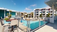 This single-level outdoor amenity space is a coastal retreat designed to maximize the beachfront lifestyle. The centerpiece is a resort-style swimming pool surrounded by lounge chairs and open deck space, enclosed by sleek metal railings that maintain unobstructed sightlines across the community. Adjacent multi-story residential buildings with modern architecture frame the pool area, complemented by mature palm trees that reinforce the Southern California beach ambiance.

A rooftop lounge area offers a refined outdoor living experience, furnished with plush white sofas accented by vibrant yellow and blue throw pillows, arranged around a built-in fire pit. From this vantage point, panoramic views of the Pacific Ocean, sandy beach, and a classic pier stretch across the horizon beneath consistently blue skies.

A signature teal-clad clubhouse pavilion anchors the deck, featuring bold black-framed glass French doors that open to the surrounding terrace. The pavilion is flanked by covered walkways with white columns, creating shaded passages ideal for year-round enjoyment. Bistro-height tables and woven chairs are scattered across the open-air terrace, providing casual gathering spots. The entire space benefits from abundant natural sunlight, coastal breezes, and a relaxed yet polished aesthetic that reflects its prime beachside setting.