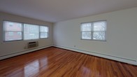 This single-floor unit offers a clean, functional layout with warm hardwood flooring that flows consistently throughout the living areas and bedroom. The open living room is generously sized, featuring two large windows that invite abundant natural light and views of the surrounding neighborhood. A wall-mounted air conditioning unit and baseboard heating ensure year-round comfort.

The kitchen is efficiently designed with rich, dark espresso-toned cabinetry, granite-style countertops, a black gas range, and a stainless steel sink — providing everything needed for everyday cooking. The layout maximizes available counter and storage space.

The bedroom continues the hardwood flooring theme and includes a built-in closet with shelving, offering practical storage solutions. A separate entry door with secure hardware connects the unit to the building's common areas.

The bathroom is finished in classic white subway tile surrounding the tub-shower combination, complemented by chrome fixtures, a white vanity with sink, and a mirrored medicine cabinet. A frosted window provides natural light while maintaining privacy.

Throughout the unit, white walls create a bright, neutral backdrop that enhances the sense of space. The overall ambiance is clean, practical, and move-in ready, with timeless finishes that suit a variety of furnishing styles.