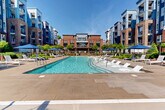 Located at 1835 Morehead Ridge Dr in Charlotte, NC, this expansive outdoor amenity area centers around a generously sized swimming pool designed for both relaxation and recreation. The pool features built-in sun shelves with in-water lounge chairs, allowing residents to enjoy the water at a shallow, comfortable depth. Surrounding the pool is a broad, well-maintained deck finished in warm-toned stone pavers, furnished with rows of white chaise lounges and large blue market umbrellas that provide ample shade throughout the day. Lush, manicured landscaping with mature trees, shrubs, and decorative plantings borders the space, creating a sense of privacy and greenery. A dedicated lounge area with a pergola and additional seating is situated adjacent to the pool, ideal for socializing or unwinding. The courtyard-style layout is framed by the multi-story residential buildings, which feature a striking combination of brick and blue paneling with private balconies overlooking the amenity space. The overall atmosphere is that of a resort-style retreat, offering a vibrant yet serene outdoor environment under the open Charlotte sky.