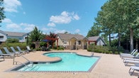 This outdoor pool area offers a well-designed recreational space centered around a freeform swimming pool with crystal-clear blue-green water. The pool features a curved, organic shape with a connected wading section, tile-trimmed edges, and clearly marked depth indicators, catering to a range of swimmers. Surrounding the pool is a generously sized stamped concrete deck with a distinctive brick-patterned finish, providing ample space for relaxation and movement around the water.

A row of modern reclining lounge chairs lines one side of the deck, ideal for sunbathing. Shaded seating is available beneath a red-painted pergola structure fitted with a large umbrella, offering a comfortable retreat from the sun. Additional pergola structures flank a stucco and stone-accented poolside building, which serves as a clubhouse or amenity facility, lending architectural character to the space.

The surrounding landscape is lush with mature trees, ornamental shrubs, and well-maintained greenery that provides natural privacy and a serene backdrop. The open sky above ensures abundant natural light throughout the day. The overall design balances functionality and aesthetic appeal, creating an inviting outdoor environment well-suited for leisure and recreation.