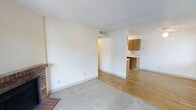 This single-floor space offers a straightforward, functional layout with a comfortable living area and an adjoining kitchen. The living room features neutral cream-colored walls, a textured ceiling, and plush carpeting that adds warmth underfoot. A classic brick fireplace anchors one corner of the room, serving as a notable focal point. A ceiling vent ensures efficient climate control throughout the space.

The living area transitions seamlessly into a kitchen area, distinguished by light hardwood-style flooring. The kitchen is equipped with warm-toned wooden cabinetry providing ample storage, along with a countertop workspace. The open sightline between the two areas contributes to a sense of connectivity and ease of movement throughout the space.

Overall, the space maintains a clean, neutral palette with white baseboards and walls that provide a versatile backdrop. The combination of carpet in the living area and hard flooring in the kitchen defines each zone clearly while maintaining a cohesive aesthetic throughout the single-floor layout.
