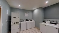 This single-floor shared laundry facility is designed with practicality and efficiency in mind. The room is finished with cool gray-toned walls and clean white tile flooring, creating a bright and tidy environment. Recessed ceiling lights provide even illumination throughout the space, ensuring a well-lit area for users at any time of day. The facility is equipped with multiple top-loading washing machines and a stacked washer-dryer unit, offering ample capacity to serve a shared community. The machines are arranged along the walls, maximizing the available floor space and allowing for comfortable movement within the room. Coin-operated controls are visible on each unit, indicating a self-service setup. An informational notice posted on the wall provides users with operating instructions and guidelines. The overall layout is straightforward and functional, with a clean, well-maintained appearance that reflects a commitment to user convenience. This laundry room serves as a reliable shared amenity, providing residents or guests with all the essentials needed for laundry in a compact and organized setting.