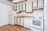 Located at 205–219 E Edgewood Blvd in Lansing, Michigan, this single-floor unit offers a practical and well-organized layout across its living spaces. The kitchen is designed for efficiency, featuring white cabinetry with warm wood-tone trim, butcher block-style countertops, a full suite of white appliances including a refrigerator, electric range, and dishwasher, and a recessed ceiling with built-in fluorescent lighting. Hardwood-style flooring runs through the kitchen and utility areas, adding warmth and easy maintenance underfoot. A standout architectural feature is the open wood shelving unit that acts as a room divider between the living area and kitchen, providing both storage and a visually distinctive separation of spaces. The living and bedroom areas are finished with plush neutral carpet and feature textured ceilings, wall-unit HVAC, and windows with vertical blinds that allow natural light to filter in. Two full bathrooms each include a tub-shower combination with clean white surrounds, and one features a single-sink vanity with storage beneath. A dedicated utility room houses an in-unit washer hookup and water heater, offering added convenience. The overall ambiance is clean, neutral, and move-in ready.