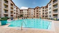 This well-appointed residential community is defined by its striking multi-story architecture, which combines neutral-toned siding with prominent natural stone accents, creating a warm and substantial aesthetic throughout the exterior. The building is arranged in a U-shaped configuration that encloses a generously sized central courtyard, making the outdoor amenity space the focal point of the community.

At the heart of the courtyard sits a large, resort-style swimming pool with clear turquoise water and a gently curved design. The surrounding pool deck is finished in neutral stone-toned pavers and is furnished with lounge chairs positioned to take full advantage of the open sky above. A covered pergola area near the poolside offers a shaded gathering spot, complemented by additional seating areas scattered across the terrace.

The courtyard grounds are thoughtfully landscaped with manicured grass areas, colorful planters, flowering shrubs, and decorative mulched garden beds, adding texture and color to the outdoor environment. Pedestrian pathways connect various sections of the courtyard, providing easy circulation throughout the space.

Individual units feature private balconies and patios with metal railings, offering residents their own outdoor retreats. Ground-floor units open to small fenced patios bordered by green lawn areas. The overall ambiance of the community is one of comfort, openness, and resort-inspired living, with the courtyard design fostering a strong sense of community in a well-maintained setting.