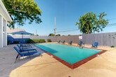Located at 1645 Carol Sue Ave in Gretna, Louisiana, this outdoor pool area offers a well-appointed recreational space designed for relaxation and leisure. The centerpiece is a generously sized rectangular swimming pool with distinctive brick coping along its perimeter, filled with clear turquoise water. Depth markers are visible, with a shallow end and a deeper section clearly indicated, and a "No Diving" designation ensures safety. The pool is accessible via built-in handrail entry points on multiple sides.

Surrounding the pool is a spacious concrete deck that provides ample room for lounging and socializing. Blue Adirondack chairs are positioned around the perimeter of the deck, offering casual seating throughout the area. On one side, a row of blue reclining lounge chairs accompanies a shaded umbrella, creating a comfortable spot to relax out of the sun. Safety equipment, including a life ring mounted to the fence, is readily accessible.

The entire area is enclosed by a tall wooden privacy fence, creating a secluded and secure environment. Mature trees rise above the fence line, providing natural shade and a touch of greenery against the open sky. The adjacent building, featuring a classic brick-and-white exterior with a covered balcony, is visible from the pool deck, adding architectural context to this outdoor amenity space.