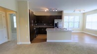 This single-floor residence showcases a thoughtfully designed open-concept layout that seamlessly connects the kitchen and living areas. The kitchen is a standout feature, appointed with rich, dark espresso-toned cabinetry that extends to the ceiling, offering generous storage. A granite-topped peninsula provides additional prep space and serves as a natural divider between the kitchen and the carpeted living area. Stainless steel appliances, including a French door refrigerator with ice and water dispenser and a range, add a sleek, modern touch. A tile backsplash complements the dark cabinetry, while track lighting overhead ensures the workspace is well-illuminated. The kitchen benefits from natural light streaming through a large window, offering views of lush greenery outside. The adjoining living space features plush, neutral-toned carpeting, creating a warm and comfortable atmosphere. The overall design balances functionality with contemporary aesthetics, making this a practical yet stylish living space.