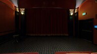 Located in the heart of Los Angeles at 1026 S Broadway, this private screening room is a masterclass in classic cinema design. The space exudes a rich, theatrical ambiance, drawing inspiration from the golden age of Hollywood with its carefully curated Art Deco aesthetic.

The room is anchored by rows of luxurious red leather seating, including individual reclining chairs and multi-seat sofas with gold piping accents, all arranged to provide optimal sightlines to the screen. A dramatic pair of deep crimson curtains trimmed with gold fringe serves as the focal point at the front of the room, framing the screen with theatrical elegance.

The walls are clad in deep burgundy tones and adorned with bold architectural elements, including tall fluted black columns topped with ornate gold-detailed capitals. Arched wall panels add a sense of grandeur, while warm wall sconces with amber-toned glass cast a moody, intimate glow throughout the space. The ceiling features a recessed coffered detail with gold trim, adding visual depth overhead.

The flooring is covered in a richly patterned dark carpet that complements the overall color palette. Subtle step lighting along the aisles ensures safe navigation in the darkened environment. Every design element in this space works cohesively to deliver an immersive, upscale cinematic experience.