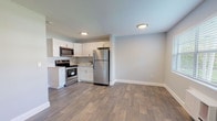 This single-floor unit showcases a clean, contemporary design with consistent wood-look tile flooring flowing throughout every room, creating a cohesive and low-maintenance living environment. The kitchen is a standout feature, equipped with stainless steel appliances including a refrigerator, electric range, and over-the-range microwave. Crisp white shaker-style cabinetry pairs with light countertops to give the kitchen a bright, modern aesthetic, while recessed lighting adds to the open and airy feel of the combined kitchen and living area. The living space is generously sized and bathed in a neutral light gray palette, offering a versatile backdrop for any furnishing style. A bedroom features a large window with horizontal blinds that frames a view of a well-maintained outdoor green space, allowing natural light to fill the room. A wall-mounted HVAC unit provides convenient climate control. The bathroom is elegantly finished with floor-to-ceiling white subway tile surrounding a soaking tub and shower combination, complete with a built-in tiled niche accented by hexagonal mosaic tile. A white vanity with brushed nickel hardware and a frameless mirror complete the polished look. Clean lines, bright finishes, and thoughtful details define this well-appointed space throughout.