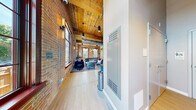 This thoughtfully designed single-floor lounge space masterfully blends industrial heritage with contemporary comfort. The open-plan layout is anchored by striking exposed brick walls that stretch upward to soaring wood-plank ceilings accented by visible structural beams and exposed ductwork — hallmarks of a beautifully preserved historic conversion. Light hardwood flooring runs throughout, providing a clean contrast to the raw materiality of the brick and timber above.

Oversized arched and grid-pane windows line multiple walls, flooding the interior with generous natural light and offering views of the surrounding greenery. The lounge area is furnished with eclectic mid-century modern seating — plush sofas, accent chairs, and ottomans in rich blues and purples — arranged around coffee tables atop colorful area rugs, creating inviting conversation zones. A wall-mounted flat-screen TV and a sleek media console anchor one side of the room.

A full-size pool table serves as a centerpiece in the gaming area, complemented by bar-height seating nearby. Reclaimed barn-style sliding doors add a rustic architectural detail that honors the building's character. A teal-accented kitchenette with open shelving, a refrigerator, and counter seating provides functional amenity. Pendant lighting and globe chandeliers add warmth, completing this dynamic and versatile communal space.