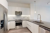 This thoughtfully designed one-bedroom, one-bathroom residence in McLean, Virginia offers a clean, contemporary aesthetic across its single-floor layout. The open-concept living and dining area is anchored by light hardwood-style flooring and large windows that invite abundant natural light and outdoor views of the surrounding neighborhood. The modern kitchen is a standout feature, showcasing flat-front white cabinetry extending to the ceiling, a marble-style subway tile backsplash, dark granite countertops, and a peninsula with an undermount sink and brushed gold faucet. Stainless steel appliances, including a range, built-in microwave, and dishwasher, complete the space. Pendant lighting and a track light fixture add a polished, contemporary touch. The bedroom is a well-proportioned retreat bathed in natural light from a wide window overlooking the exterior. It connects directly to the full bathroom, which features large-format tile flooring, a soaking tub with marble surround, a floating vanity with granite countertop, and a framed mirror. Two closets provide generous storage, including one outfitted with a wire shelving organization system. An in-unit laundry area adds everyday convenience to this well-appointed McLean residence.