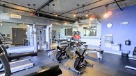 This fitness facility offers a comprehensive workout environment designed to accommodate a wide range of exercise routines. The space features an industrial-inspired aesthetic, with exposed ductwork and piping running across the ceiling, complemented by pendant lighting that provides a warm, focused ambiance throughout the room.

The gym is outfitted with multiple stationary cycling bikes positioned prominently on durable rubber flooring, ideal for high-intensity cardio sessions. A well-stocked dumbbell rack lines one side of the space, offering a variety of weights for strength training. A multi-functional cable machine is positioned nearby, expanding the options for resistance-based exercises.

Large wall-mounted mirrors span a significant portion of one wall, enhancing the sense of space and allowing users to monitor their form during workouts. Stability balls and additional accessories are neatly stored within reach. A window along the far wall allows natural light to filter into the space, offering a connection to the outdoors. The rubber-tiled flooring provides both safety and comfort underfoot, making the gym a practical and well-appointed facility for fitness enthusiasts.