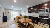 This single-floor residence offers a thoughtfully designed open-concept layout that seamlessly connects the kitchen, dining, and living areas. The kitchen is outfitted with rich dark espresso cabinetry, granite countertops, stainless steel appliances including a refrigerator, range, and built-in microwave, all set above durable light wood-style flooring. A round wooden dining table with industrial-style chairs provides an inviting space for meals, positioned conveniently between the kitchen and living area. The living room features a comfortable sectional sofa, warm neutral tones, and direct access to a private balcony through sliding glass doors that allow abundant natural light to fill the space. The bedroom is a calm retreat, appointed with plush carpeting, neutral walls, and ample room for furniture. The full bathroom includes a granite-topped dark vanity, a tiled shower-tub combination, and matching wood-style flooring that ties the space together. A utility area houses the water heater and includes wire shelving for additional storage. Recessed lighting throughout the main living areas enhances the bright, modern ambiance of this well-designed residence.