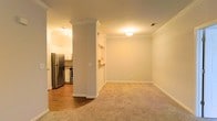 This single-floor space offers an open, functional layout that seamlessly connects the living area to the kitchen. The living room is finished with wall-to-wall carpeting in a neutral tone, creating a warm and comfortable atmosphere. Crown molding runs along the ceiling throughout, adding a refined architectural detail that elevates the overall aesthetic. A decorative column with crown molding accents serves as a subtle divider between the living space and the kitchen area, providing definition without closing off the open feel.

The kitchen is accessible via a pass-through opening in the wall, allowing for easy interaction between the two spaces. It features hardwood flooring, white cabinetry, stainless steel appliances including a refrigerator and range, and stone-look countertops, giving it a clean and modern appearance. Recessed and flush-mount ceiling lighting throughout ensures both spaces are well-illuminated. The neutral color palette of warm beige tones on the walls and trim contributes to a cohesive, inviting ambiance across the entire space.