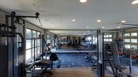 This fitness center is thoughtfully designed to support a comprehensive workout experience. The space is anchored by a combination of durable flooring — including rubberized lifting areas and patterned carpet tiles — that segments zones for different types of training. Large multi-pane windows line the walls, flooding the interior with natural light and offering views of the surrounding greenery, creating an open and energizing atmosphere. Recessed lighting throughout the ceiling ensures consistent illumination during any hour of the day. Floor-to-ceiling mirrors span a significant portion of the walls, visually expanding the space while allowing users to monitor their form during exercise. The equipment selection is extensive, featuring cable and pulley machines, a dedicated free weights and bench press station, a dumbbell rack, medicine balls, and a row of cardio machines including treadmills along the far wall. The ceiling height contributes to a spacious, airy feel that prevents the space from feeling crowded. Overall, the fitness center strikes a balance between functional design and an inviting environment, making it well-suited for a broad range of fitness routines from strength training to cardiovascular conditioning.
