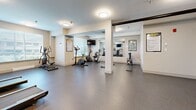 This fitness center offers a thoughtfully organized single-level workout space designed to accommodate a variety of exercise needs. The room is finished with durable, speckled rubberized flooring throughout, providing a practical and resilient surface suited for both cardio and strength training activities.

The cardio section features multiple treadmills and stationary bikes, including upright and recumbent models, positioned for comfortable use. A cable resistance machine and a functional adjustable bench serve the strength training area, complemented by a stability ball and a dumbbell rack visible in the mirrored section of the room. Large wall-mounted mirrors along one side visually expand the space and allow users to monitor their form during workouts.

Multiple flat-screen televisions are mounted at strategic points throughout the room, ensuring visibility from various workout stations. Exercise guide charts and informational posters are displayed on the walls, adding a functional and educational element to the space. Flush-mount ceiling lights provide consistent, even illumination throughout the room. Structural columns divide the space into distinct workout zones while maintaining an open, accessible flow. A window along one wall introduces natural light, contributing to the room's clean, neutral aesthetic.