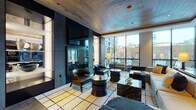 Located in Chicago's West Loop neighborhood, this single-floor amenity space at 34 N Sangamon Street delivers a refined, contemporary atmosphere designed for both relaxation and social gatherings. The lounge area anchors the space with a striking two-sided glass fireplace set within a dark stone surround, framed by illuminated display shelving and topped with a wall-mounted television. A wood-paneled ceiling adds warmth and texture overhead, while large floor-to-ceiling windows flood the interior with natural light and offer views of an outdoor pool area. Generously scaled seating arrangements feature neutral-toned sofas, leather lounge chairs, and sculptural black side tables atop a boldly patterned area rug. A dedicated entry foyer greets visitors with large-format tile flooring and a dramatic hand-painted mural spanning an entire wall, establishing an artistic tone from arrival. The kitchen and dining area is equally well-appointed, featuring dark cabinetry with stone accents, stainless steel appliances, and a long communal dining table illuminated by globe pendant lights. Built-in shelving and intimate booth-style seating provide additional gathering spots throughout, completing a space that balances style with functionality.