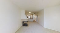 This single-floor space features an open-concept layout that connects the living area to the kitchen, creating a functional and cohesive flow throughout. The living area is finished with neutral-toned carpet and is anchored by a corner glass-faced fireplace, which serves as a notable architectural focal point. The fireplace's transparent design adds visual depth and warmth to the space.

The kitchen is positioned toward the rear and is accessible via a raised breakfast bar that provides both a natural division and an additional seating or workspace surface. The kitchen features light cabinetry, a dark countertop, and includes a refrigerator. A window above the sink area allows natural light to filter into the cooking space.

The overall color palette is consistently neutral, with white walls and light-toned finishes throughout, contributing to a clean and airy atmosphere. A hallway visible beyond the kitchen suggests additional rooms or utility areas. The ceiling height is standard, and flush-mounted lighting provides uniform illumination. The open design and cohesive materials give the space a practical yet comfortable residential character.