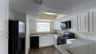 The kitchen is the focal point of this single-floor space, designed with a practical U-shaped layout that maximizes efficiency and storage. White shaker-style cabinetry lines the walls, providing ample storage space above and below the countertops, which feature a neutral gray laminate surface. Black appliances, including an electric range, built-in microwave, dishwasher, and refrigerator, create a cohesive contrast against the white cabinetry. A double-basin sink is positioned beneath a window, allowing natural light to filter into the space and offering a pleasant view while working. Above the sink area, a recessed tray ceiling with an integrated skylight panel adds an architectural detail that enhances the sense of openness and introduces additional overhead lighting. The wood-style laminate flooring throughout the kitchen provides durability and a warm visual element. A textured ceiling adds dimension overhead, while a central HVAC vent ensures climate control. The overall layout is clean and functional, designed to support everyday use with straightforward, low-maintenance finishes throughout.
