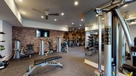 This spacious fitness center is thoughtfully designed to accommodate a full range of workout needs within a single, open-plan floor. The space is divided into distinct functional zones that flow seamlessly into one another. Exposed brick accent walls run throughout, lending an industrial-chic aesthetic that is complemented by smooth white ceilings, recessed lighting, and ceiling fans that maintain airflow across the entire facility.

The cardio zone features a long row of treadmills and elliptical machines positioned along mirrored walls, which visually expand the space and allow users to monitor their form. An adjacent cycling area is outfitted with stationary bikes equipped with individual digital screens, alongside wall-mounted flat-screen televisions for on-demand workout content. Natural light filters in through large windows in this section, offering views of an outdoor area.

The strength training zone includes a comprehensive selection of selectorized machines, adjustable benches, and a well-stocked dumbbell rack with kettlebells and free weights of varying sizes. Floor-to-ceiling mirrors line this area as well, maximizing visibility and spatial openness. Rubber-composite flooring covers the entire facility, providing durability and impact absorption throughout.