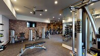 This spacious fitness center is thoughtfully designed to accommodate a full range of workout needs within a single, open-plan floor. The space is divided into distinct functional zones that flow seamlessly into one another. Exposed brick accent walls run throughout, lending an industrial-chic aesthetic that is complemented by smooth white ceilings, recessed lighting, and ceiling fans that maintain airflow across the entire facility.

The cardio zone features a long row of treadmills and elliptical machines positioned along mirrored walls, which visually expand the space and allow users to monitor their form. An adjacent cycling area is outfitted with stationary bikes equipped with individual digital screens, alongside wall-mounted flat-screen televisions for on-demand workout content. Natural light filters in through large windows in this section, offering views of an outdoor area.

The strength training zone includes a comprehensive selection of selectorized machines, adjustable benches, and a well-stocked dumbbell rack with kettlebells and free weights of varying sizes. Floor-to-ceiling mirrors line this area as well, maximizing visibility and spatial openness. Rubber-composite flooring covers the entire facility, providing durability and impact absorption throughout.