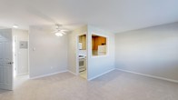 This single-floor residential space offers a clean, functional layout designed for comfortable everyday living. The living area is bright and open, featuring soft carpeted flooring, neutral light-toned walls, and a ceiling fan that adds both practicality and a sense of airiness to the room. The space is well-lit with a calm, understated ambiance that makes it easy to personalize.

A standout architectural detail is the pass-through window cut into the wall between the living area and the kitchen, creating a sense of openness and visual connectivity between the two spaces. This design element allows light and sightlines to flow freely, making the overall footprint feel more expansive.

The kitchen is accessible through a doorway adjacent to the pass-through and is equipped with warm honey-toned wood cabinetry, a white gas range with oven, a refrigerator, and a microwave, providing all the essential appliances for a fully functional cooking space. The kitchen's layout is compact yet efficient, with cabinetry extending to the upper walls for ample storage.

Overall, the space presents a tidy, move-in-ready environment with a cohesive neutral color palette throughout, making it a practical and inviting single-floor residence.