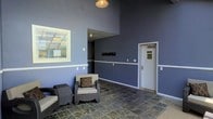 This single-floor space functions as a welcoming lounge or lobby area, distinguished by its notably high ceilings that lend an open, airy feel to the interior. The walls are painted in a two-tone scheme, with a soft periwinkle-blue upper section transitioning to a lighter hue below a crisp white chair rail, creating a visually structured yet relaxed atmosphere.

The flooring consists of large slate-style tiles in muted earth tones, adding a grounded, textural contrast to the cool wall palette. A pendant light fixture with a warm-toned bulb hangs from the ceiling, providing a focal point overhead while contributing to the overall ambient lighting.

The seating area features wicker-framed chairs with cream-colored cushions and patterned decorative pillows, arranged to encourage casual conversation. A framed artwork depicting an architectural or exterior scene is mounted on the left wall, adding visual interest.

A recessed wall niche with mounted hooks provides functional storage or display space. A standard door on the right wall leads to an adjacent area, suggesting this space serves as a transitional or communal gathering zone within a larger facility.