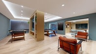 This well-appointed single-floor amenity space offers a generously sized, open layout designed for both social interaction and relaxed recreation. The interior is unified by light-toned hardwood flooring that runs throughout, complementing the muted teal-blue accent walls and crisp white ceilings outfitted with recessed and track lighting. A central built-in wood shelving unit with integrated cabinetry acts as a stylish room divider, separating functional zones while maintaining an open, connected feel. The main game area is anchored by a solid walnut ping-pong table positioned beneath a tray ceiling with ambient LED cove lighting, adding depth and a refined finish to the space. A wall-mounted flat-screen television, framed within a light wood surround, serves as a media focal point alongside the game area. An adjacent lounge zone features bold rust-orange upholstered armchairs with black metal frames, arranged against a decorative feature wall adorned with a gold geometric pattern on a deep blue background. A separate, glass-enclosed media lounge provides a quieter retreat, furnished with a sofa and additional television. Curated artwork accents the walls throughout, contributing to the space's modern, design-forward aesthetic. A foosball table is also present, further enhancing the recreational offerings of this thoughtfully designed amenity space.