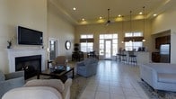 This single-floor space offers an open, multi-functional layout designed for comfort and community gathering. The great room immediately impresses with its soaring ceilings, which lend a sense of grandeur and openness to the entire area. A classic white-mantled fireplace anchors one side of the room, providing a warm focal point complemented by plush lounge seating, including cushioned armchairs and a sofa arranged around a dark wood coffee table.

The space transitions seamlessly into a dining area, where a table and chairs are positioned to take advantage of the natural light streaming through large windows dressed with warm-toned roman shades. French doors lead to the exterior, further enhancing the bright and airy atmosphere while offering easy outdoor access.

A bar-height counter with sleek metal stools defines a casual seating nook near the windows, ideal for informal conversation or light refreshments. Recessed lighting throughout the ceiling, along with stylish pendant lights and a ceiling fan, ensure the space is well-lit at any hour. Light-toned tile flooring runs throughout, adding to the clean, open feel of the interior. Together, these features create a welcoming and versatile space suited for relaxation and social gatherings alike.