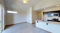 This single-story space showcases a bright, open-concept design that seamlessly connects the living area and kitchen. The living space is highlighted by vaulted ceilings that enhance the sense of volume, complemented by a ceiling fan with integrated lighting. A clerestory window near the roofline allows natural light to filter into the space, contributing to its airy feel. The flooring throughout consists of light-toned laminate or luxury vinyl planks, lending a clean and contemporary aesthetic. The walls are finished in a neutral off-white tone, providing a versatile backdrop. The kitchen features a white peninsula-style island with an integrated sink and a built-in overhang for bar seating. Dark-toned upper cabinetry provides contrast against the lighter finishes, and recessed lighting illuminates the kitchen area. A hallway visible beyond the kitchen suggests additional rooms further into the unit. The overall design emphasizes an open, minimalist aesthetic with practical features, making efficient use of the available space while maintaining a modern and functional layout throughout.