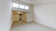 This single-floor space showcases a striking open layout defined by its notably high ceilings that create an expansive, airy atmosphere. A row of clerestory windows positioned near the top of the wall allows natural light to pour into the main living area, brightening the neutral, light-toned walls and contributing to the space's open and welcoming feel.

The main living area features wall-to-wall carpeting in a neutral tone, providing a comfortable and cohesive foundation throughout. A lowered ceiling section transitions into an adjacent area, which houses a flush-mount ceiling light and leads toward the kitchen, where white appliances are visible. This architectural split-level ceiling detail adds visual interest and helps define the separate functional zones within the space.

A utility or laundry area is also accessible from the interior, indicated by visible mechanical components. A door on the far wall of the main living area provides an additional point of entry or access to a secondary space. HVAC vents are integrated throughout, ensuring year-round comfort. The overall design is clean and minimal, with a neutral color palette of whites and soft beiges that lends itself well to a variety of interior styles and furnishing choices.