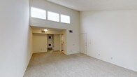 This single-floor space showcases a striking open layout defined by its notably high ceilings that create an expansive, airy atmosphere. A row of clerestory windows positioned near the top of the wall allows natural light to pour into the main living area, brightening the neutral, light-toned walls and contributing to the space's open and welcoming feel.

The main living area features wall-to-wall carpeting in a neutral tone, providing a comfortable and cohesive foundation throughout. A lowered ceiling section transitions into an adjacent area, which houses a flush-mount ceiling light and leads toward the kitchen, where white appliances are visible. This architectural split-level ceiling detail adds visual interest and helps define the separate functional zones within the space.

A utility or laundry area is also accessible from the interior, indicated by visible mechanical components. A door on the far wall of the main living area provides an additional point of entry or access to a secondary space. HVAC vents are integrated throughout, ensuring year-round comfort. The overall design is clean and minimal, with a neutral color palette of whites and soft beiges that lends itself well to a variety of interior styles and furnishing choices.