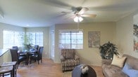 This single-story space offers an open, connected layout that integrates the living and dining areas into one cohesive environment. The living area is anchored by warm hardwood flooring that extends throughout, contributing to an inviting and unified aesthetic. A ceiling fan with integrated lighting is centrally positioned overhead, providing both airflow and ambient illumination.

Multiple windows fitted with horizontal blinds allow generous natural light to filter into the space, creating a bright and airy atmosphere. The neutral, sage-toned walls serve as a versatile backdrop that complements a range of furnishing styles. A patterned accent chair adds texture and visual interest to the seating area, which is further defined by a round, dark-finished coffee table set atop an area rug.

To the side, the dining area accommodates a table and chairs, benefiting from additional window views of surrounding greenery. Decorative wall art and potted plants enhance the lived-in warmth of the space. The overall layout is practical and well-proportioned, offering comfortable, functional living within a modest and well-maintained footprint.