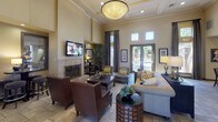 This single-floor communal space presents a welcoming and well-appointed interior designed for comfort and social gathering. The entryway sets a refined tone with decorative wall niches displaying tasteful accents, stone-tiled flooring, and glass-paneled doors that draw natural light into the foyer.

The main living area is expansive, characterized by soaring ceilings with crown molding and a large statement pendant light fixture. A central fireplace with an ornate iron screen serves as a focal point, flanked by a wall-mounted television and framed artwork. Seating is arranged in multiple clusters, combining leather armchairs, upholstered sofas, and accent chairs to accommodate various group sizes. Stone tile flooring extends throughout, complementing the warm, neutral color palette.

A bar-height kitchen area adjoins the lounge, featuring a granite-topped island with barstool seating, pendant lighting, upper cabinetry with a tile backsplash, and stainless steel appliances. High ceilings continue into this zone, reinforcing the open, airy atmosphere.

Glass block windows and full-length glazed doors connect the interior to outdoor views, enhancing the brightness and inviting ambiance throughout the space.