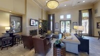 This single-floor communal space presents a welcoming and well-appointed interior designed for comfort and social gathering. The entryway sets a refined tone with decorative wall niches displaying tasteful accents, stone-tiled flooring, and glass-paneled doors that draw natural light into the foyer.

The main living area is expansive, characterized by soaring ceilings with crown molding and a large statement pendant light fixture. A central fireplace with an ornate iron screen serves as a focal point, flanked by a wall-mounted television and framed artwork. Seating is arranged in multiple clusters, combining leather armchairs, upholstered sofas, and accent chairs to accommodate various group sizes. Stone tile flooring extends throughout, complementing the warm, neutral color palette.

A bar-height kitchen area adjoins the lounge, featuring a granite-topped island with barstool seating, pendant lighting, upper cabinetry with a tile backsplash, and stainless steel appliances. High ceilings continue into this zone, reinforcing the open, airy atmosphere.

Glass block windows and full-length glazed doors connect the interior to outdoor views, enhancing the brightness and inviting ambiance throughout the space.