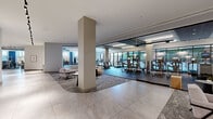 This single-floor fitness facility is thoughtfully designed to deliver a comprehensive wellness experience within a refined, contemporary setting. The space is organized into several distinct zones, each serving a specific purpose while maintaining a cohesive aesthetic throughout.

The cardio area is generously proportioned, housing rows of modern treadmills, elliptical trainers, and upright bikes equipped with individual display screens. Floor-to-ceiling windows line the perimeter, allowing abundant natural light and outdoor views to energize the space. Rubber sport flooring, slatted wood ceiling accents, and linear LED lighting contribute to a polished yet functional atmosphere.

A dedicated free-weight and strength training zone features rubber flooring, large wall-mounted mirrors, organized dumbbell racks, stability balls, and various cable and resistance machines, providing ample room for functional movement and strength work.

Separating the fitness areas from the rest of the facility are full-height glass partitions with sleek black hardware, creating visual openness while defining each zone clearly. Adjacent to the workout spaces is a stylish lounge and transition area finished with large-format stone-look tile flooring, curated artwork on white walls, and comfortable seating arranged around a wood-accented nook. The overall ambiance is clean, upscale, and welcoming.