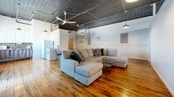 Located at 179 St Paul St in the heart of Rochester, NY, this urban loft blends industrial character with comfortable living across a single spacious floor. Rich, wide-plank hardwood floors run throughout, complemented by striking ornate tin ceilings that lend a historic, industrial aesthetic to every room. The open-concept living area is generously proportioned, anchored by exposed ceiling pipes, pendant lighting, and a ceiling fan, creating an airy, loft-like ambiance. A kitchen with essential appliances is seamlessly integrated into the living space. The primary bedroom is a standout, offering ample room, large windows dressed with layered, colorful curtains that admit natural light, and the same eye-catching tin ceiling detail overhead. The second bedroom provides a comfortable, versatile space with hardwood floors, white-painted brick walls, and exposed industrial elements. The bathroom continues the loft's aesthetic with white-painted brick walls, hardwood flooring, a window for natural light, and a shower with curtain enclosure. Throughout the unit, white walls and bright natural light from multiple windows balance the bold industrial elements, creating a space that feels both characterful and inviting.