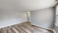 This 1-bedroom, 1-bathroom unit offers a thoughtfully arranged layout across a single floor, accessible via a carpeted staircase with white-painted railings. The main living area features smooth, light gray walls, clean white baseboards, and durable wood-look vinyl plank flooring that lends a modern, cohesive feel. A baseboard heating system runs along the perimeter, ensuring efficient climate control throughout. The kitchen is equipped with granite-style countertops, white cabinetry, a black electric range, and an over-the-range microwave, complemented by a subway tile backsplash and a bright window that brings in natural light. The bedroom is a generously sized room finished with plush gray carpeting, light gray walls, and a multi-pane window that allows for natural light. Baseboard heating is also present, adding to the room's comfort. A full bathroom rounds out the unit, featuring white tile surrounding a tub-shower combination, a white vanity with a single-basin sink, and light tile flooring — all maintaining the clean, neutral palette found throughout the space. Overall, the unit presents a consistent, move-in-ready aesthetic with a neutral color scheme and practical finishes throughout.
