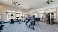 Located in Denton, TX, this fitness center offers a comprehensive exercise environment across a single, open floor. The space is thoughtfully designed with high ceilings and large, multi-pane windows lining the walls, allowing abundant natural light to fill the room and providing views of the surrounding outdoor areas, including a pool. Large ceiling fans provide additional airflow and comfort throughout the space.

The exercise room is carpeted in a blue-toned, speckled carpet that spans the entire floor. It is equipped with a wide variety of fitness equipment, including treadmills, elliptical machines, a stair climber, and a stationary bike to accommodate cardiovascular training. A multi-functional cable machine and a heavy punching bag serve strength and functional training needs, while a dumbbell rack rounds out the free weight options.

Full-length mirrors with gold-toned frames are mounted along one wall, visually expanding the space and allowing users to monitor their form during workouts. Built-in wood storage benches and cabinetry provide practical storage solutions. Wall-mounted flat-screen televisions and framed artwork add to the polished, modern aesthetic. An interactive fitness mirror is also mounted on the wall, offering guided workout capabilities. The overall ambiance is bright, modern, and motivating.