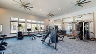 Located in Denton, TX, this fitness center offers a comprehensive exercise environment across a single, open floor. The space is thoughtfully designed with high ceilings and large, multi-pane windows lining the walls, allowing abundant natural light to fill the room and providing views of the surrounding outdoor areas, including a pool. Large ceiling fans provide additional airflow and comfort throughout the space.

The exercise room is carpeted in a blue-toned, speckled carpet that spans the entire floor. It is equipped with a wide variety of fitness equipment, including treadmills, elliptical machines, a stair climber, and a stationary bike to accommodate cardiovascular training. A multi-functional cable machine and a heavy punching bag serve strength and functional training needs, while a dumbbell rack rounds out the free weight options.

Full-length mirrors with gold-toned frames are mounted along one wall, visually expanding the space and allowing users to monitor their form during workouts. Built-in wood storage benches and cabinetry provide practical storage solutions. Wall-mounted flat-screen televisions and framed artwork add to the polished, modern aesthetic. An interactive fitness mirror is also mounted on the wall, offering guided workout capabilities. The overall ambiance is bright, modern, and motivating.