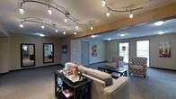 This single-floor space functions as a well-appointed lounge or common area, designed to offer a comfortable and inviting environment. The room is illuminated by a striking curved track lighting system mounted on a textured ceiling, providing bright, directional light across the space. Complementary flush-mount ceiling fixtures extend illumination into an adjacent seating alcove, ensuring the entire area remains well-lit.

The main seating area features a neutral-toned sofa paired with dark wood side and coffee tables, creating a casual yet organized gathering space. Decorative items displayed on the tables add a personalized touch. A large framed mirror on one wall enhances the sense of openness and reflects natural light.

Beyond the primary seating area, a secondary lounge zone is furnished with patterned armchairs and a glass-topped coffee table, positioned near a window that allows natural light to filter in. A decorative art piece adds a pop of color to the neutral wall tones. Dark baseboards and carpeted flooring run throughout, contributing to the cohesive, grounded aesthetic of the space.