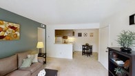 Located at 848 Tamarack Dr in Fayetteville, North Carolina, this single-floor residence offers a practical and comfortable layout designed for easy living. The open-concept main area seamlessly connects the living room, dining space, and kitchen, creating a cohesive and functional floor plan. The living room features neutral-toned carpeting and a warm accent wall in sage green, adding a touch of color contrast to the otherwise light and airy interior. A textured ceiling runs throughout, complementing the clean, simple aesthetic of the space. The half-wall pass-through kitchen is equipped with dark wood upper cabinetry, a white range, refrigerator, and built-in overhead ventilation, providing efficient use of the space while maintaining an open feel to the main living area. Adjacent to the kitchen, the dining area comfortably accommodates a bistro-style table and seating, making it well-suited for casual meals. A sliding closet door is visible along one wall, offering convenient built-in storage. Natural light filters into the space, contributing to a bright and welcoming atmosphere throughout. The overall design prioritizes functionality and comfort, making it a practical single-floor living option in Fayetteville.