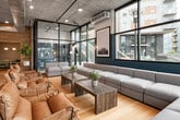 This contemporary single-floor commercial space showcases an industrial-modern aesthetic defined by exposed concrete ceilings, warm light hardwood flooring, and expansive floor-to-ceiling black-framed windows that flood the interior with natural light and offer sweeping views of the surrounding urban streetscape. The open main area is thoughtfully zoned to accommodate both collaborative and focused work, featuring a generously sized modular lounge with neutral upholstered sectional seating, rustic wooden coffee tables, and accent leather chairs arranged around large-format framed artwork mounted against deep teal feature walls. A statement dining-style conference table with ornate iron legs anchors a dedicated meeting zone, complemented by sleek bar-height stools. Glass-partitioned private meeting rooms with frosted lower panels and teal interior accents provide semi-private spaces for focused gatherings, while recessed alcoves lined with warm wood-slat wall paneling offer intimate nook seating for smaller conversations. The space also includes a fully accessible restroom finished with navy blue subway tile wainscoting, hexagonal floor tile, and stainless steel grab bars. Pendant lighting, industrial track fixtures, and abundant glazing throughout contribute to the space's open, airy, and professionally inviting ambiance.