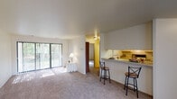 This single-floor residence is designed with a practical, open layout that maximizes the flow between living spaces. The main living area is generously proportioned, finished with neutral-toned carpet underfoot and bright white walls that create a clean, airy atmosphere. A full-width sliding glass door fitted with vertical blinds fills the space with natural light and provides a view of surrounding greenery.

The kitchen is efficiently arranged in a galley configuration, featuring white cabinetry with wood-trimmed accents, white countertops, a full-size refrigerator, and an electric range. A pass-through bar counter opens directly to the living area, accommodating bar-height seating and enhancing the connected feel of the space.

The bedroom is a well-sized, carpeted room with neutral walls and a window that allows for natural light. The full bathroom includes a tiled tub-shower combination with built-in shelving, a white vanity with storage, and tile flooring. Throughout the unit, the consistent use of white and warm neutral tones contributes to a cohesive, understated aesthetic well-suited to a variety of furnishing styles.