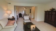 This single-story space is designed with an open-concept layout that seamlessly connects the living and dining areas, creating a cohesive and functional flow. The living area is carpeted and furnished with a sofa, accent chair, and a dark-toned coffee table, establishing a comfortable and inviting atmosphere. The neutral walls and light-colored carpet contribute to a clean, understated aesthetic throughout.

A wide arched passageway connects the living space to the dining area, which features a round dining table with chairs and is accented by a ceiling fan with a light fixture overhead — a practical detail that enhances air circulation and ambient lighting. Wall art and decorative pieces add modest character to the space.

Adjacent to the dining area, a bar-height counter with stools suggests a kitchen nearby, offering an informal dining or prep space. A hallway visible in the background leads to additional rooms, providing a sense of depth and privacy beyond the main living areas. Overall, the space presents a straightforward, livable layout with consistent finishes and a calm, neutral ambiance throughout.