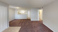 This single-floor residence features a practical, well-organized layout with distinct living zones connected by an open floor plan. The living area is generously sized, finished with dark wall-to-wall carpeting, neutral gray walls, and a textured ceiling. A large window allows natural light to fill the space, offering views of the surrounding exterior. The living area flows seamlessly into the kitchen, which is separated by a raised breakfast bar that doubles as a convenient partition. The kitchen follows a galley-style configuration, equipped with white cabinetry, light-toned countertops, a stainless steel sink, and a stainless steel electric range with an overhead ventilation hood. Tile flooring provides a clean, durable surface underfoot in this area. The bedrooms continue the carpeted aesthetic, maintaining a consistent, neutral color palette with ample wall space and vertical-blind-covered windows that allow adjustable natural light. The bathrooms are finished with tile flooring and feature full tub-shower combinations with chrome fixtures, a toilet, and a vanity with countertop space. Clean white finishes throughout the bathrooms contribute to a tidy, functional feel. Overall, this space offers a cohesive and straightforward design suited for comfortable everyday living.