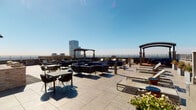 This rooftop amenity level offers a well-appointed outdoor experience high above the surrounding cityscape. The open-air terrace is finished with large-format stone tiles and features an array of seating configurations, including modular lounge suites with navy cushions, dining tables with contemporary woven chairs, and sun loungers arranged along the perimeter. Wooden pergolas punctuate the space, providing defined gathering zones and partial shade across the terrace. Potted greenery and flowering plants add natural color throughout.

A full-size swimming pool is positioned along one side of the rooftop, enclosed by expansive floor-to-ceiling glass walls and doors that allow abundant natural light to fill the interior pool area while maintaining unobstructed views of the skyline beyond. The pool deck extends outward, connecting seamlessly to the open terrace.

Sturdy metal railings line the edges of the terrace, ensuring safety while preserving panoramic views of the urban skyline, which includes prominent high-rise towers and extends toward the horizon. The brick building exterior adds an architectural warmth that contrasts with the sleek tile finishes underfoot. Whether used for social gatherings, leisure, or exercise, this rooftop amenity space delivers a versatile and visually striking environment elevated above the city.