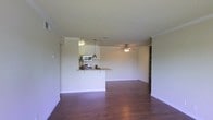 This single-floor residence offers an open-concept layout that seamlessly blends the kitchen and living areas into one cohesive, functional space. The rich, dark hardwood flooring runs throughout, adding warmth and a polished aesthetic to the interior. A textured ceiling with crown molding lends a classic finishing touch overhead, while a ceiling fan in the living area ensures year-round comfort. The kitchen is defined by a raised peninsula counter that serves as a natural divider between the cooking and living zones, providing both prep space and an informal dining area. White cabinetry lines the kitchen walls, complemented by pendant lighting that casts a warm, focused glow over the counter. Stainless steel appliances, including a microwave and range, are neatly integrated into the cabinetry. A built-in shelving unit on the far wall of the living area offers practical display and storage options. The open layout and neutral color palette create a bright, airy ambiance throughout, making the space feel welcoming and versatile. This well-appointed single-level home is designed for both comfortable everyday living and easy entertaining.