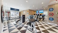 Located in Woodbury, Minnesota, this fitness center offers a comprehensive and thoughtfully designed exercise space that caters to a wide range of workout preferences. The room is immediately striking thanks to its bold black-and-white geometric triangle-patterned flooring, which sets an energetic tone throughout the space.

Cardio equipment includes treadmills and stationary cycling bikes, each equipped with individual screens for interactive workout experiences. A large wall-mounted television provides additional entertainment for those using the machines. Strength training is well-represented with a full dumbbell rack, weight benches, and resistance machines from Life Fitness, accommodating both beginners and seasoned athletes.

A standout feature is the red MoveStrong functional training tower, which supports a variety of bodyweight and cable-based exercises and serves as a bold visual centerpiece. Colorful weight plates are displayed on the surrounding walls, adding a lively, organized aesthetic. Floor-to-ceiling glass partitions along one side of the room offer views into an adjacent lounge area and allow natural light to filter in, creating an open, connected atmosphere. Large windows near the exit doors further enhance the brightness of the space. Recessed ceiling lighting ensures the room remains well-lit throughout. The overall design balances function and style, delivering a modern, motivating environment for fitness.