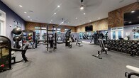 This fitness center offers a generously sized, single-floor exercise space designed to support a comprehensive workout experience. The room features a sophisticated aesthetic, with deep navy-blue walls accented by striking geometric wood-panel wall treatments that lend warmth and texture to the space. Recessed lighting and ceiling fans provide a well-lit, comfortable environment throughout.

The facility is thoughtfully divided into distinct training zones. A dedicated cardio area lines one side of the room, featuring a row of treadmills, elliptical machines, and other aerobic equipment, all positioned near large windows that invite natural light and offer views of the surrounding neighborhood. A separate strength training zone accommodates a full cable machine, a Smith machine or squat rack, multiple adjustable benches, and an organized dumbbell rack with a wide range of weights. Wall-mounted mirrors expand the visual depth of the space and support proper form during workouts.

Large flat-screen televisions are mounted prominently for entertainment during workouts. Durable rubber flooring throughout provides safety and comfort underfoot. Bright glass-paneled entry doors enhance the openness of the space. Overall, the fitness center presents a clean, modern, and well-appointed environment suited for a full range of cardio and strength training activities.