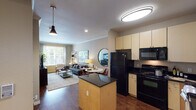 This single-floor residence offers a thoughtfully designed open-concept layout that seamlessly connects the kitchen, dining, and living areas. The kitchen is equipped with black appliances, including a full-size refrigerator, gas range, and built-in microwave, all complemented by light maple cabinetry and dark granite countertops. A central island with matching granite provides additional prep space and casual seating. Rich hardwood-style flooring runs throughout the main living areas, adding warmth and continuity to the space.

The living room is bright and welcoming, benefiting from large windows that invite generous natural light and offer views of the surrounding greenery. Crown molding along the ceiling lends an elegant architectural detail to the space. A leaning ladder shelf serves as both a functional and decorative element, while the open floor plan allows for flexible furniture arrangements.

The bedroom is carpeted for comfort and features a ceiling fan, a large window with blinds and curtains, and ample space for storage and display shelving. The full bathroom is well-appointed with a dark granite vanity, light maple cabinetry, a large wall mirror with accent lighting, and a shower-tub combination. Overall, the unit presents a cohesive and functional living environment with consistent finishes throughout.